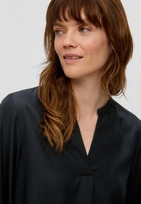 Schwarze Bluse mit Stehkragen, glatter Textur und langen Ärmeln. Verfügt über eine dezente vordere Falte und ein minimalistisches Design.