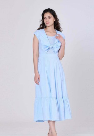 Femme aux cheveux bouclés portant une robe midi sans manches bleu clair avec un nœud noué au niveau de la poitrine, debout devant un fond uni.