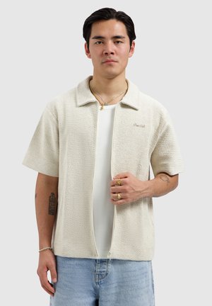 Jonge man die een open, crèmekleurig textuur kortärmlig overhemd draagt over een wit shirt, lichtblauwe spijkerbroek en gouden sieraden tegen een effen achtergrond.