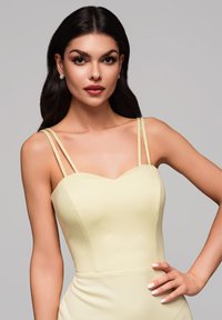 Robe jaune ajustée avec un décolleté en cœur, des bretelles fines et une texture lisse. Le mannequin a les cheveux droits et porte des boucles d'oreilles en studs.