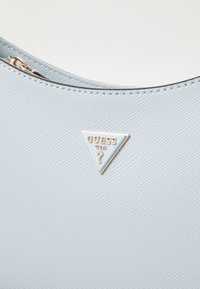 Sac en cuir vegan texturé bleu clair avec un logo en métal doré en forme de triangle portant l'inscription "GUESS USA". Fermeture éclair lisse en haut.