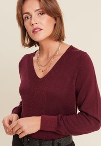 Bordeaux sweater met een V-hals en lange mouwen. Beschikt over een textuur centraal paneel en geribbelde manchetten, gedragen met gelaagde gouden kettingen.