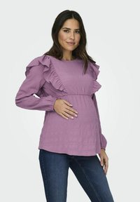 Lavanda majčinska bluza s volanima, teksturiranom tkaninom i nabranim strukom. Uparena s tamnim trapericama, nosi je model.