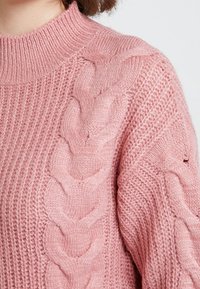 Pull rose en tricot avec un col montant, un motif torsadé le long de la manche et une texture côtelée sur le corps.