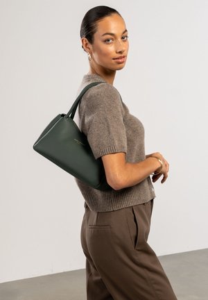 Sac à main en cuir vert au design structuré, avec une anse courte et un logo discret, porté sur un haut en tricot beige et un pantalon marron.