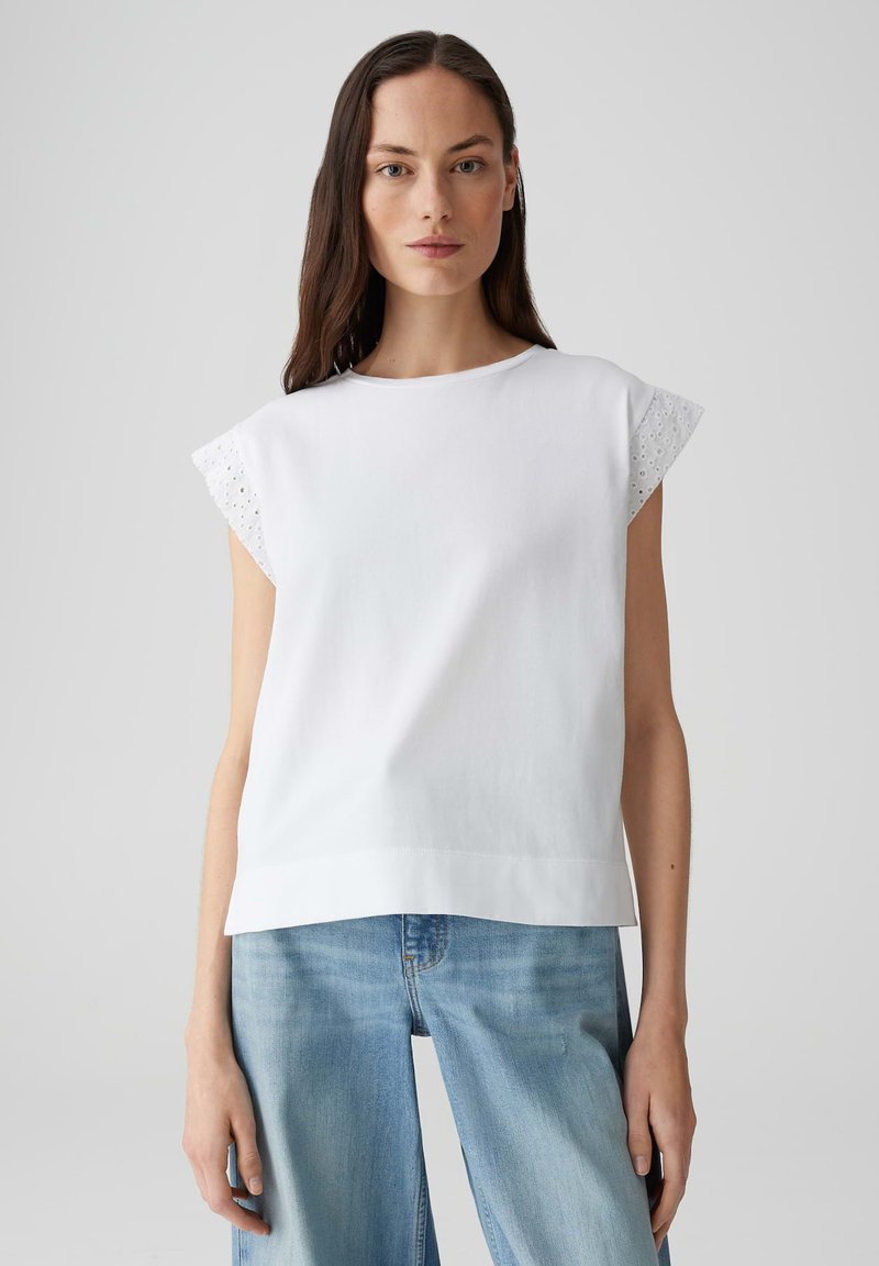 Witte katoenen top met korte uitlopende mouwen en een oogjesafwerking. Rechte silouet, ronde halslijn en een gladde textuur. Gecombineerd met blauwe jeans.