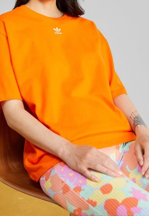 Osoba ubrana w jaskrawą pomarańczową koszulkę Adidas i kolorowe kwiatowe spodnie, siedząca na brązowym krześle, pokazująca tatuaż na lewym przedramieniu.