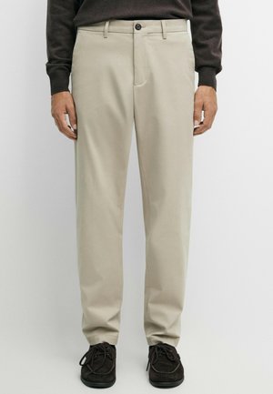 Homme portant un pantalon beige à jambes droites, des chaussures noires à lacets et un haut sombre à manches longues, debout devant un fond uni.