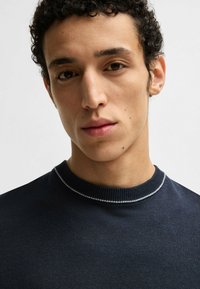 Dark blue knit sweater with a textured finish and a light gray ribbed collar. Close-up of a young man’s face, showcasing a neutral expression.