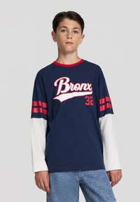 Granatowa koszulka z czerwonymi akcentami, z logo "Bronx" i numerem "32". Widoczne długie białe rękawy. Wykonana z bawełny, o luźnym kroju.