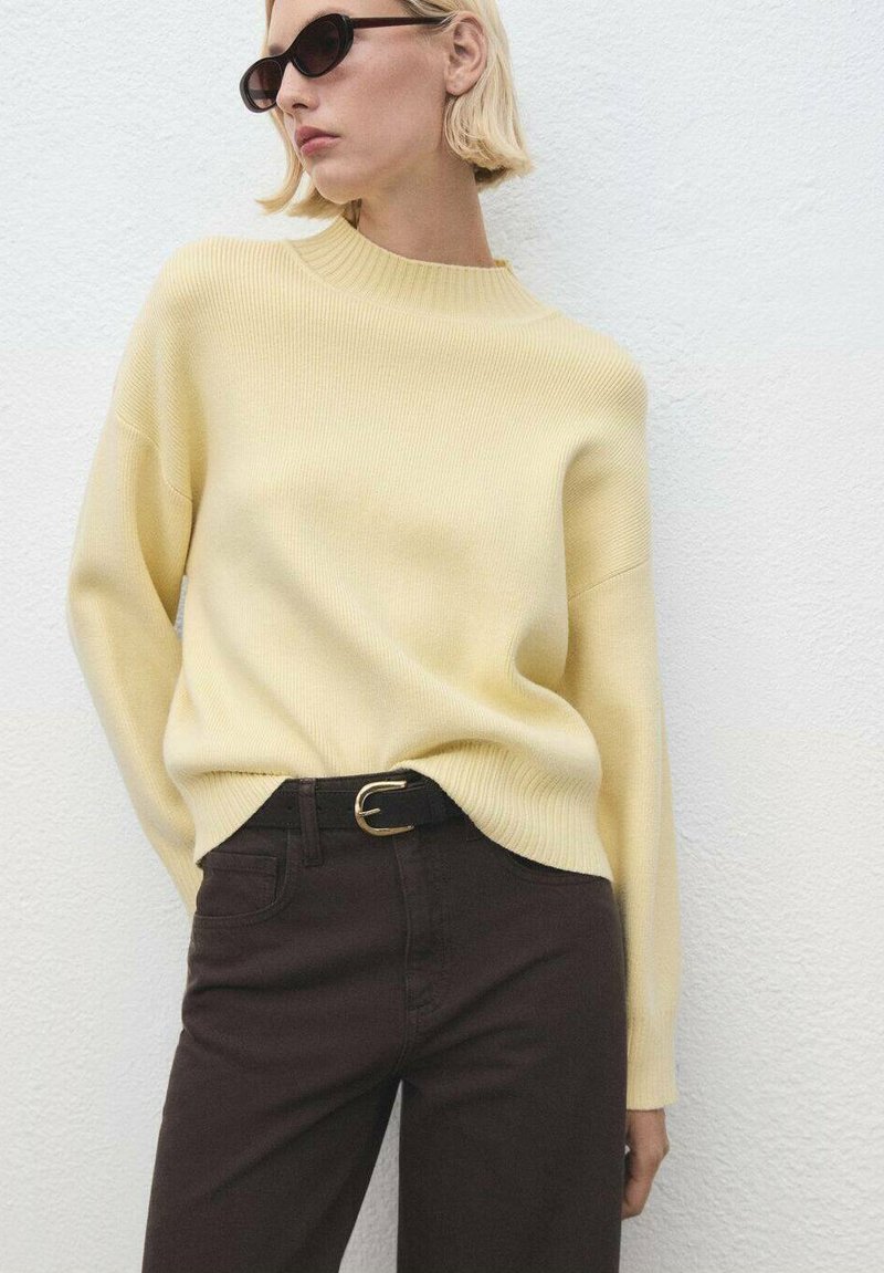 Suéter amarillo con canalé y cuello alto, combinado con jeans marrón oscure. El conjunto incluye un cinturón negro con una hebilla circular dorada.