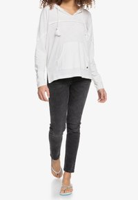 Sudadera con capucha gris claro, con un bolsillo frontal, cuerdas ajustables y puños acanalados, combinada con jeans ajustados negros y chanclas.