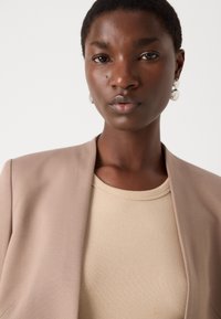 Beige blazer over a light beige ribbed shirt; smooth fabric, tailored design, with subtle sheen. Large silver earrings accentuate the look.