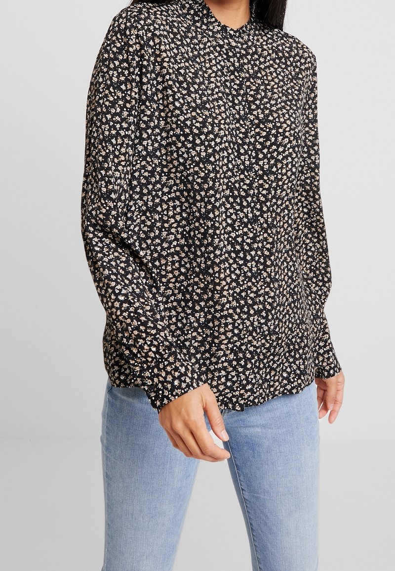 Personne portant un chemisier noir à manches longues avec un petit imprimé floral blanc et beige, associé à un jean bleu clair, debout devant un fond uni.