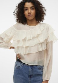 Blouse crème à volants avec manches longues, en tissu transparent, multiple couches de volants sur le devant, associée à un jean en denim taille haute.