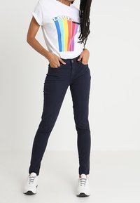 Camiseta blanca con gráfico de arcoíris "LOVE", pantalones ajustados navy y zapatillas blancas. El conjunto presenta un diseño casual y contemporáneo.