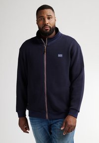Marinblå zip-up sweatshirt med en texturerad mönster, ståkrage och elastiska muddars. Har en märkespatch på övre vänstra sidan. Bärs med blå jeans.