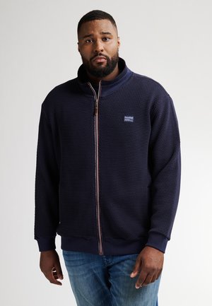 Felpa navy con zip, caratterizzata da un motivo testurizzato, colletto rialzato e polsini elasticizzati. Presenta un'etichetta con il marchio sulla parte superiore sinistra. Indossata con jeans blu.