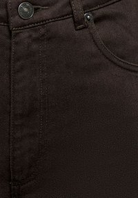 Gros plan sur un jean en denim noir montrant le bouton, la passoire de ceinture, la poche avant et les détails de couture.