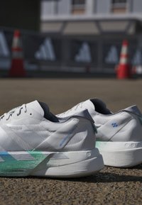Witte hardloopschoenen met blauwe en groene accenten geplaatst op buitenasfalt, met wazige verkeerskegels en reclamebanners op de achtergrond.