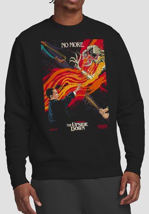 Man wearing black sweatshirt with colorful Stranger Things Upside Down artwork showing a person using psychic power against a monster, others with weapons.
