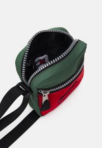 Borsa compatta con tessuto verde e rosso, apertura con zip, interno nero, tasca frontale, tracolla regolabile e dettagli in metallo argentato.