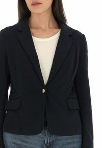Blazer blu navy in tessuto texture, chiusura a un bottone, revers con incisione e due tasche frontali, indossato sopra una maglietta bianca e jeans blu.