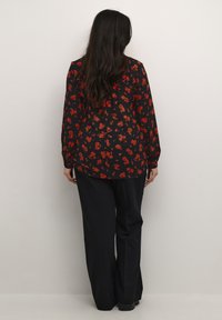 Sort blomsterbluse med røde blomster, lange ærmer og en løs pasform; sammen med sorte vide bukser. Ensfarvet baggrund.