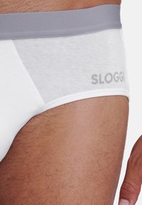 Weiße Baumwollunterhose mit einem grauen elastischen Bund, auf dem der Markenname "SLOGGI" in Grau steht. Glatte Textur, tief sitzendes Design.