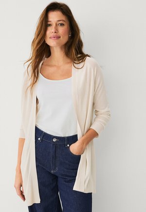Hellbeige Strickjacke aus weichem Stoff, mit einem lässigen, offenen Frontdesign, langen Ärmeln und glatter Textur. Kombiniert mit einem weißen Oberteil und dunklen Jeans.