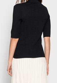 Svart ribbad stickad topp med en åtsittande silhuett och korta ärmar, med en krage och textilstruktur. Bärs med en plisserad vit kjol.