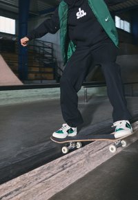 Skateboarder in a green vest and black hoodie performs a trick on a skateboard. Shoes feature white, green, and black colors with contrasting accents.
