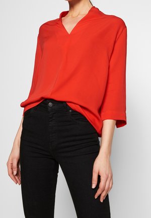Femme portant un chemisier rouge ample à col en V avec manches trois-quarts et pantalon noir taille haute ajusté sur un fond uni.