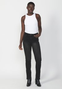 Top blanco sin mangas combinado con unos jeans skinny negros de cintura alta. La modelo lleva botas de tacón alto negras. Colores sólidos y diseño ajustado.