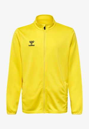 Dreng med blonde dreadlocks iført en lysegul sportsjakke og shorts, sorte sokker og sorte sneakers, stående med den ene hånd i lommen.