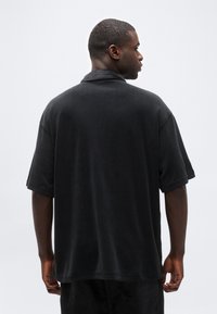 Άνδρας που φοράει φαρδιά μαύρη κοντομάνικη πουκάμισο με γιακά, στέκεται με την πλάτη και το κεφάλι γυρισμένα δεξιά, μπροστά σε απλό λευκό φόντο.