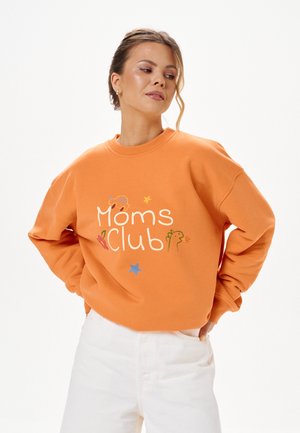 Woman with light brown hair wearing an orange sweatshirt with "Moms club" text and small colorful drawings, paired with white pants.