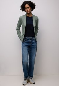 Femme aux cheveux bouclés portant un blazer sauge texturé, une chemise bleu marine, un jean bleu et des baskets blanches, debout les mains dans le dos.