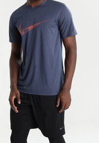 Mörkblå atletisk skjorta i lätt tyg, med en röd Nike swoosh-logotyp på bröstet, tillsammans med svarta shorts.