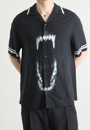 Osoba ubrana w czarną koszulę na guziki z krótkim rękawem, z białymi paskami na rękawach i dużą grafiką otwartych szczęk zwierzęcia z ostrymi zębami.