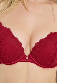 Reggiseno push-up in pizzo rosso con bordi smerlati, caratterizzato da una fascia liscia in raso e un piccolo dettaglio metallico al centro. Design strutturato su tutta la superficie.
