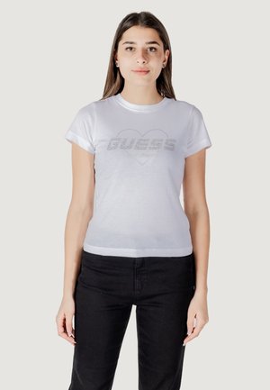 T-shirt blanc à manches courtes avec un logo "GUESS" pailleté intégré dans une forme de cœur. Le tissu est lisse et ajusté, associé à un pantalon noir.