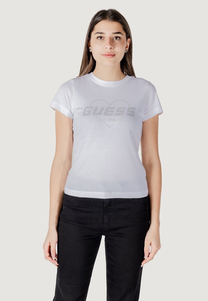 Witte t-shirt met korte mouwen met een glinsterend "GUESS" logo in de vorm van een hart. De stof is glad en aansluitend, gecombineerd met zwarte broek.