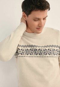 Kremowy sweter z granatowym geometrycznym wzorem na piersi, gładka tekstura, ściągacze przy mankietach i dekolcie. Dopasowany krój z długimi rękawami.