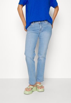 Personne portant un jean bleu clair, un t-shirt bleu vif et des tongs à plateforme vert clair, debout les mains dans les poches.