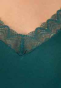 Top in tessuto teal con delicata rifinitura in pizzo lungo l'escollatura a V su pelle chiara.
