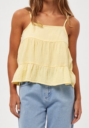 Femme portant un haut jaune sans manches à volants, associé à un jean en denim bleu clair, debout devant un fond uni.