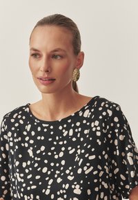 Fekete felső nagy krémszínű pettyekkel; puha anyag, bő szabás, kerek nyakkivágás. A modell texturált arany ovális fülbevalót visel.