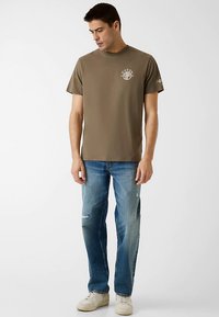 Olivgrünes T-Shirt mit kurzen Ärmeln und einem grafischen Logo auf der Brust, kombiniert mit hellblauen, verwaschenen Jeans und cremefarbenen Sneakers.