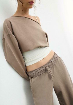 Mlada žena u bež dukserici s otkrivenim ramenima, bijelom kratkom majicom i odgovarajućim bež trenirkom s vezicama na svijetloj pozadini.